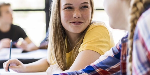 Female friends talking in classroom. Confident girls are sitting at table in high school. Students are wearing casuals.